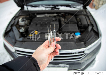 Modern ceramic glow plugs in hand against the background of a diesel car engine in winter in cold weather. Breakdown and replacement of glow plug, close-up Modern ceramic glow plugs in hand against the background of a diesel car engine in winter in cold weather. Breakdown and replacement of glow plug, close-up 113025253