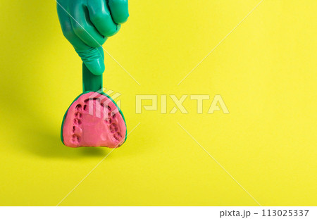 The hand of a dentist doctor in a green medical glove holds a dental spoon with an alginate plastic mass for making a plaster cast of the patient s jaw on a yellow background. Concept of malocclusion, The hand of a dentist doctor in a green medical glove holds a dental spoon with an alginate plastic mass for making a plaster cast of the patient s jaw on a yellow background. Concept of malocclusion, 113025337