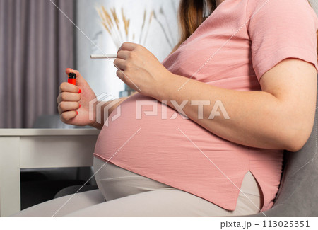 A pregnant girl with a big belly sits in the kitchen at the table and smokes. Smoking and pregnancy. Harm to the health of the child and mother. 113025351