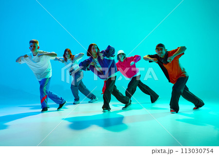 Group of talented dancers in retro clothes practices choreographed number in neon light against gradient studio background. 113030754