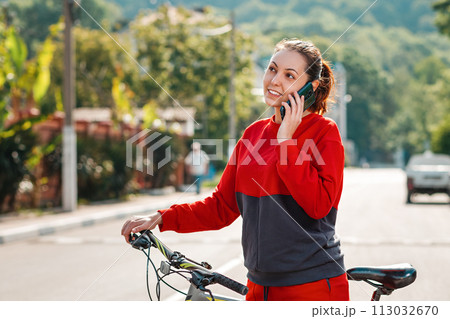 Communication. A portrait of young beautiful smiling woman is making a phone call and pushing a Bicycle. Sunny street on the background. Outdoor. Modern technology 113032670