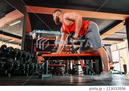 A young bearded athletic man trains with dumbbells in the gym. Bottom view. The concept of a sporty lifestyle and fitness A young bearded athletic man trains with dumbbells in the gym. Bottom view. The concept of a sporty lifestyle and fitness 113032686