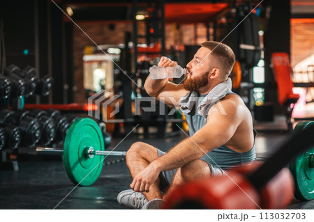 A young bearded athletic man trains in the gym. Man drinking a bottle of water. The concept of fitness and training A young bearded athletic man trains in the gym. Man drinking a bottle of water. The concept of fitness and training 113032703