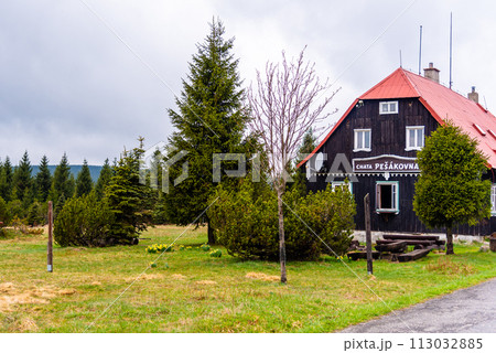 The Pesakovna wooden cottage stands surrounded by lush greenery and coniferous trees in the serene Jizerka village, under a cloudy sky. The Pesakovna wooden cottage stands surrounded by lush greenery and coniferous trees in the serene Jizerka village, under a cloudy sky. 113032885