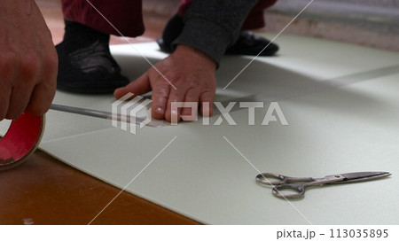 male hands glue together with transparent tape sheets of soft light-colored underlay for laminate on the floor and scissors lying nearby, do it yourself preparing the flooring at home male hands glue together with transparent tape sheets of soft light-colored underlay for laminate on the floor and scissors lying nearby, do it yourself preparing the flooring at home 113035895