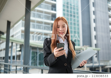 A young Asian businesswomen wearing a suit holding files standing in a big city on a busy downtown street. Young Asian businesswoman using smartphone texting to contact clients. 113036533