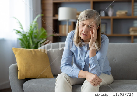 An elderly woman appears distressed and alone, sitting pensively on a couch in a well-lit living room, evoking emotions of loneliness and concern. 113037724