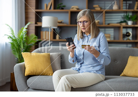 Anxious mature woman sitting on sofa, holding credit card and phone, looking at screen with concern, suspecting a fraudulent transaction. 113037733