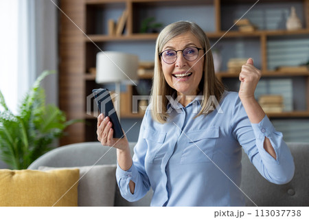 Elderly woman holding a smartphone in a casual indoor setting appears elated or triumphant, possibly celebrating good news. 113037738