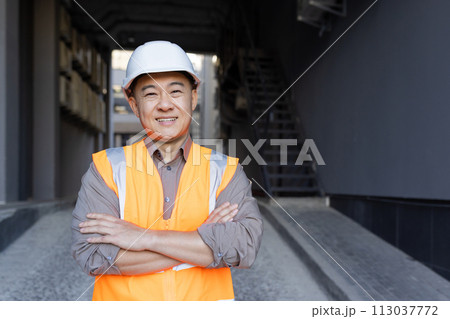 Close-up portrait of an Asian young man in a construction helmet and vest, an employee of an engineering firm, an architect standing in front of the camera smiling and crossing his arms over his chest Close-up portrait of an Asian young man in a construction helmet and vest, an employee of an engineering firm, an architect standing in front of the camera smiling and crossing his arms over his chest 113037772