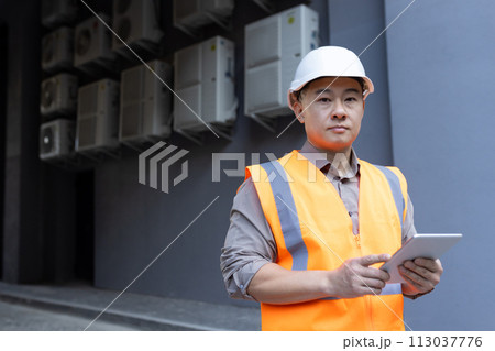 Close-up portrait of a young Asian man wearing a hard hat and vest, standing outside a factory, construction site, holding a tablet and looking seriously into the camera. 113037776