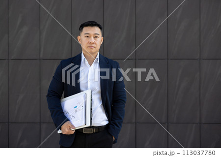 Portrait of a young successful serious young Asian man standing outside a business center against a wall, holding documents and an agreement in his hand. He looks seriously into the camera. 113037780