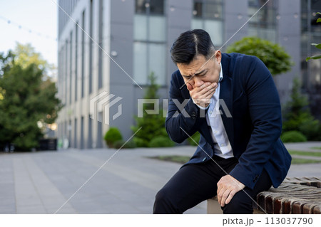 Asian young man in a suit sits on a bench in the street with his hand covering his mouth, feels severe nausea and urge to vomit. 113037790