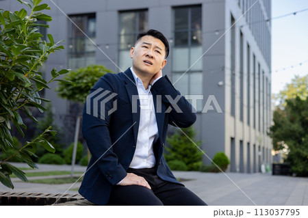 Outside the office building, a young Asian man in a suit is sitting on a bench, holding his hand by his neck, tired from work, doing a massage, feeling pain in his muscles. 113037795