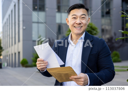 Portrait of smiling young businessman man standing outside office, holding documents and open envelope, received good news, looks happily at camera. Portrait of smiling young businessman man standing outside office, holding documents and open envelope, received good news, looks happily at camera. 113037806