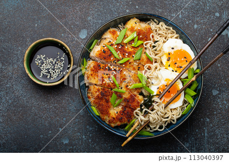 Asian noodles ramen soup with deep fried panko chicken fillet and boiled eggs in ceramic bowl with chop sticks, soy sauce on stone rustic background top view 113040397