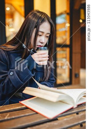 カフェの外の席で飲み物を飲みながら本を読む女子高生 113043986