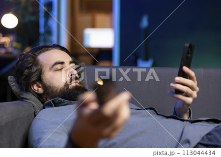 Bored person lounged on couch binging series on streaming services, browsing using remote and texting on phone. Man watching VOD shows on television, zapping though channels, chatting on smartphone 113044434