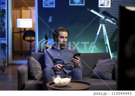 Person in home theater binging series on streaming service, texting friend on phone. Man in neon lit apartment with bowl of popcorn in front watching shows on television and chatting with mate 113044443
