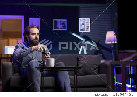 Gamer immersed in virtual world, holding controller in modern apartment, playing videogames. Cheerful caucasian man enjoys gaming session from the comfort of his living room 113044450