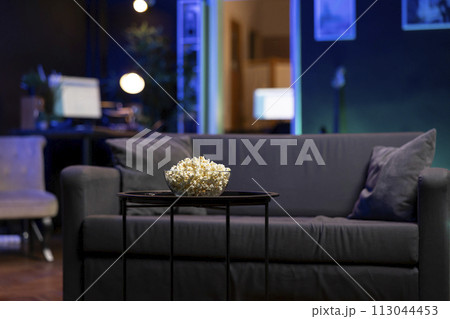 Focus on bowl of popcorn on table with neon illuminated home theater apartment in blurry background. Salty snack prepared for upcoming movie night in cozy RGB lit living room 113044453