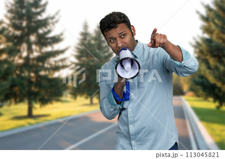 Young indian man talking into megaphone and pointing with index finger. Man with loudspeaker standing outdoors. 113045821