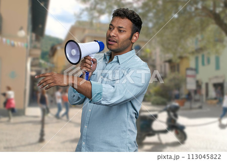 Young man with megaphone standing on blur city street background. Indian man with loudspeaker outdoors. Young man with megaphone standing on blur city street background. Indian man with loudspeaker outdoors. 113045822