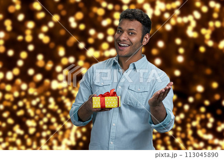 Delighted young man showing gift box and looking at camera. Festive bokeh background. Best Christmas gift shop. 113045890