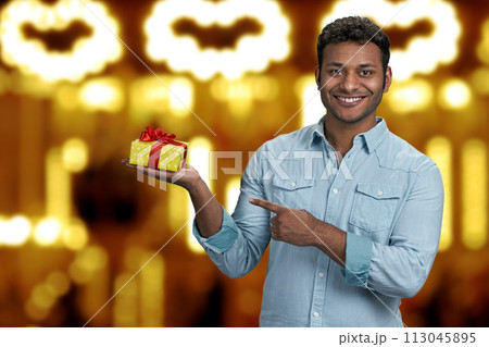 Cheerful young man showing gift box in his hand. Golden bokeh lights background. Christmas holiday sales. 113045895