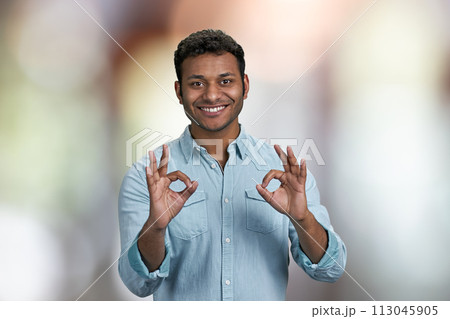 Positive smiling young man showing okey gesture with both hands. Handsome guy gesturing okey on abstract bokeh background. 113045905