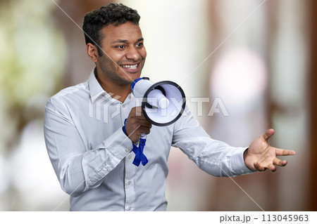 Cheerful young man in white shirt talking into megaphone. Abstract bokeh background. 113045963