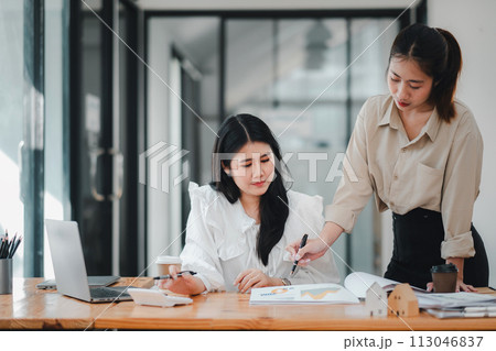 Two female real estate professionals discuss project details with a model house on the table, symbolizing project planning and teamwork. 113046837