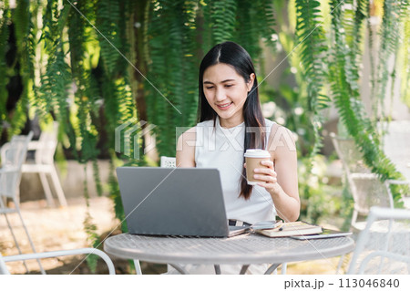 Happy freelancer works on her laptop while holding a coffee cup, sitting at a garden table surrounded by verdant plants. Happy freelancer works on her laptop while holding a coffee cup, sitting at a garden table surrounded by verdant plants. 113046840