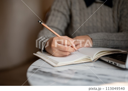 A young Asian female student is sitting indoors and writing on her notebook with a pencil. A young Asian female student is sitting indoors and writing on her notebook with a pencil. 113049540