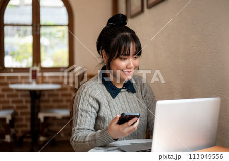 A smiling, positive young Asian woman is working remotely at a coffee shop, working on her laptop. 113049556