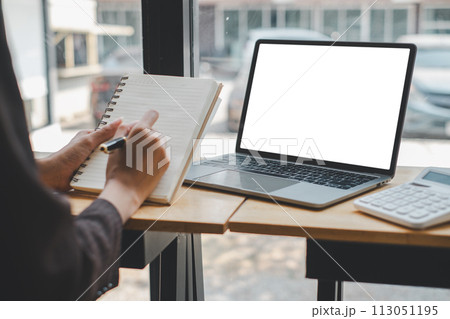 Person is diligently writing in a notebook beside a sleek laptop, with a calculator at hand, indicating a focus on business planning, budgeting, or financial analysis in a bright, modern workspace. 113051195