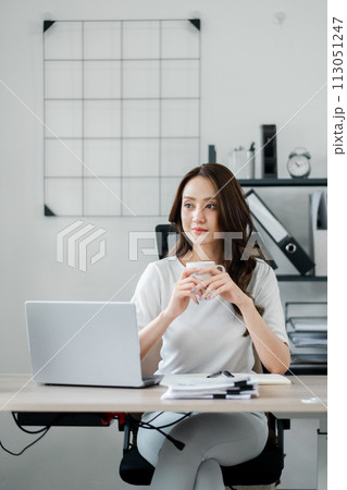 Contemplative businesswoman holding a mug, deep in thought at her minimalist home office, with a laptop and paperwork on her desk. 113051247