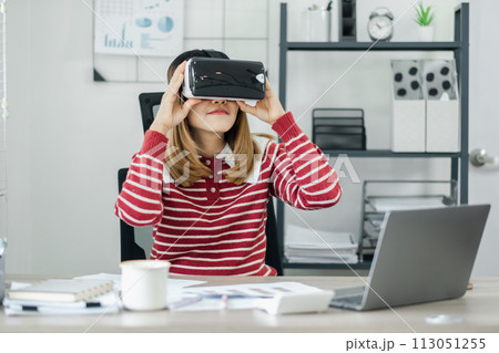 Woman with blonde hair in a striped sweater adjusts her virtual reality headset at an office desk with graphs on the wall. Woman with blonde hair in a striped sweater adjusts her virtual reality headset at an office desk with graphs on the wall. 113051255
