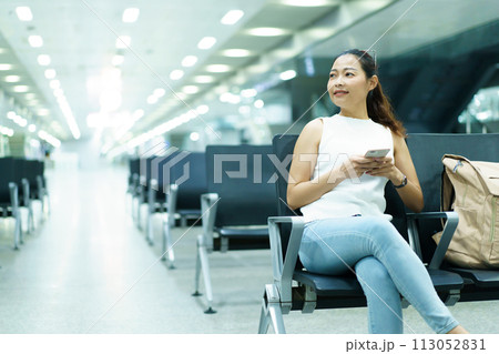 Happy beautiful Asian woman passenger sitting in airport departure terminal alone and sends a message or chats with her friends. Solo female traveler waiting for boarding at the gate in airport. 113052831