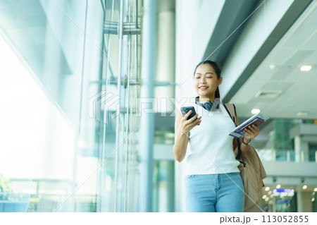 Happy beautiful Asian solo female tourist walking in the airport terminal, woman walking and talking with her friend on mobile phone. Asian woman passenger walks in airport terminal to departure gate. 113052855