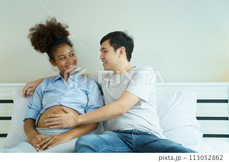 Happy cheerful husband and pregnant wife staying - relaxing together in bedroom, husband embracing or playing on pregnant wife tummy. Diverse ethnicity husband and wife lifestyles concept. 113052862