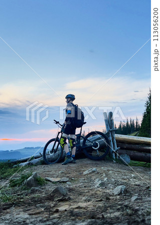 Cyclist man riding electric bike outdoors in the evening. Male tourist resting on hill, enjoying beautiful mountain landscape, wearing helmet and backpack. Concept of active leisure. Cyclist man riding electric bike outdoors in the evening. Male tourist resting on hill, enjoying beautiful mountain landscape, wearing helmet and backpack. Concept of active leisure. 113056200