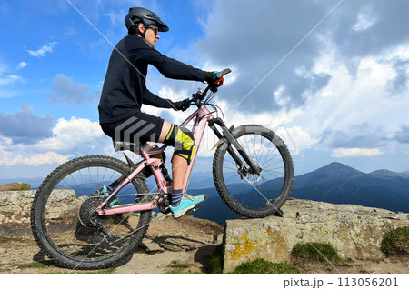 Cyclist man riding electric bike outdoors on sunny day. Male tourist resting on grassy hill, enjoying beautiful mountain landscape, wearing helmet. Concept of active leisure. 113056201