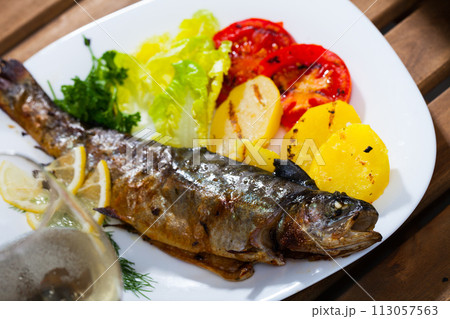 Baked whole trout with potatoes, tomatoes and greens on plate, glass of wine 113057563