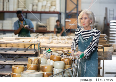 Elderly worker in ceramics factory carries many different clay ceramic cups and plates in cart 113057659