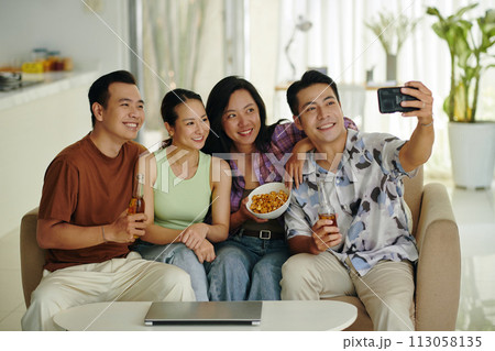 Group of smiling friends sitting on couch at home and taking selfie 113058135