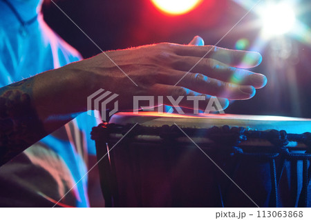 Close-up of male hands, musician playing bongo drums on dark background with stage lights. African culture live event 113063868