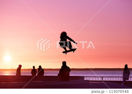 Unrecognizable teenage boy silhouette showing high jump tricks on scooter against 113070548