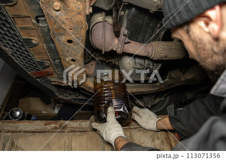 Black motor oil is drained under the car in a pit in the garage. Car maintenance in the garage with engine oil change. The engine oil is drained into a jar to be replaced with new oil. Black motor oil is drained under the car in a pit in the garage. Car maintenance in the garage with engine oil change. The engine oil is drained into a jar to be replaced with new oil. 113071356