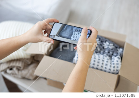 Natural wool sweaters are folded in a box with a recycling sign for reuse. A housewife takes pictures of folded clothes with her phone camera and places an advertisement for sale on the Internet. Natural wool sweaters are folded in a box with a recycling sign for reuse. A housewife takes pictures of folded clothes with her phone camera and places an advertisement for sale on the Internet. 113071389